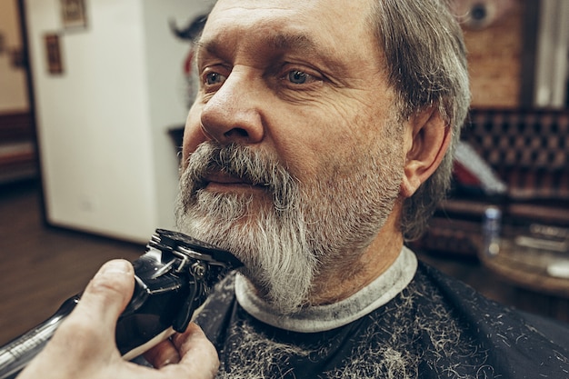 Close-up side view portrait of handsome senior bearded caucasian man getting beard grooming in modern barbershop.