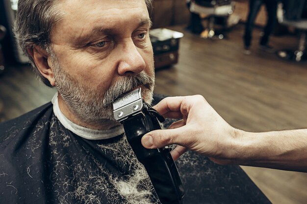 Close-up side view portrait of handsome senior bearded caucasian man getting beard grooming in modern barbershop.