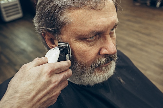 Close-up side view portrait of handsome senior bearded caucasian man getting beard grooming in modern barbershop.