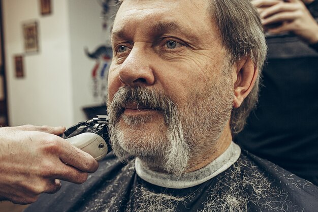 Close-up side view portrait of handsome senior bearded caucasian man getting beard grooming in modern barbershop.