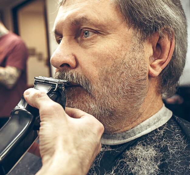 Close-up side view portrait of handsome senior bearded caucasian man getting beard grooming in modern barbershop.