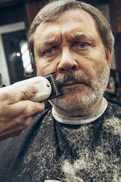 Close-up side view portrait of handsome senior bearded caucasian man getting beard grooming in modern barbershop.