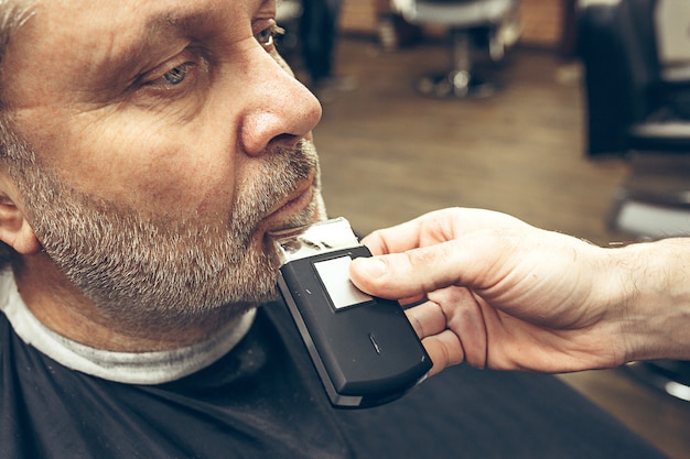 Free photo close-up side view portrait of handsome senior bearded caucasian man getting beard grooming in modern barbershop.