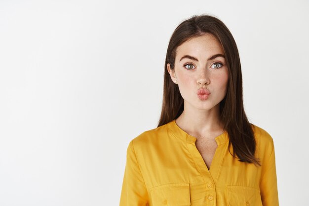 Close-up of silly and cute girl pucker lips, making kissing shape and looking at camera, standing over white wall.