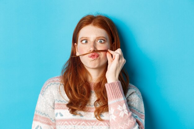 Close-up of silly and funny redhead girl making moustache with hair strand and puckered lips, grimacing against blue background