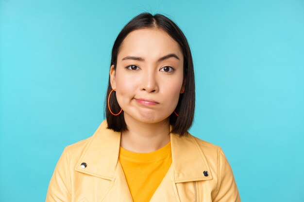 Close up of skeptical asian girl looking disappointed staring with doubt or disbelief grimacing at camera standing over blue background