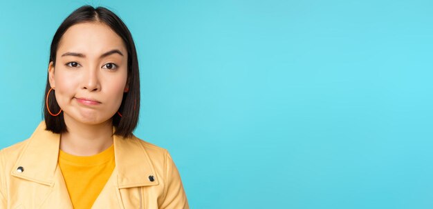 Close up of skeptical asian girl looking disappointed staring with doubt or disbelief grimacing at camera standing over blue background