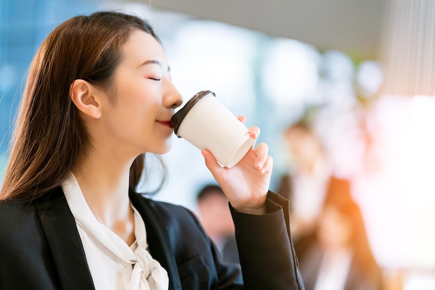 Крупным планом умная привлекательная азиатская деловая женщина улыбается рукой держать горячий напиток с современным офисным фоном