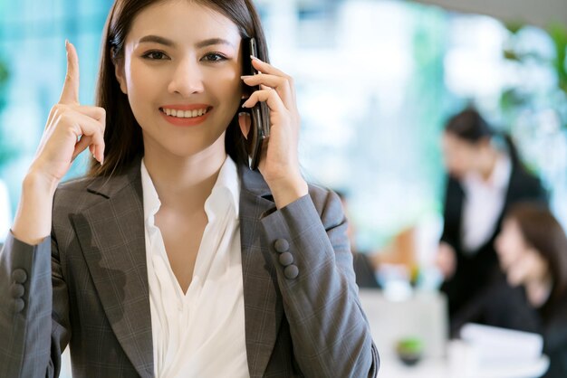 스마트한 매력적인 아시아 비즈니스 여성 미소 손을 잡고 현대적인 사무실 배경으로 스마트폰을 닫습니다