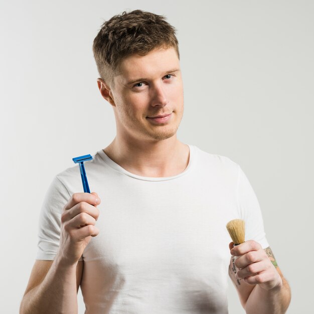 Close-up of smart young man holding razor and shaving brush in hands against white backdrop