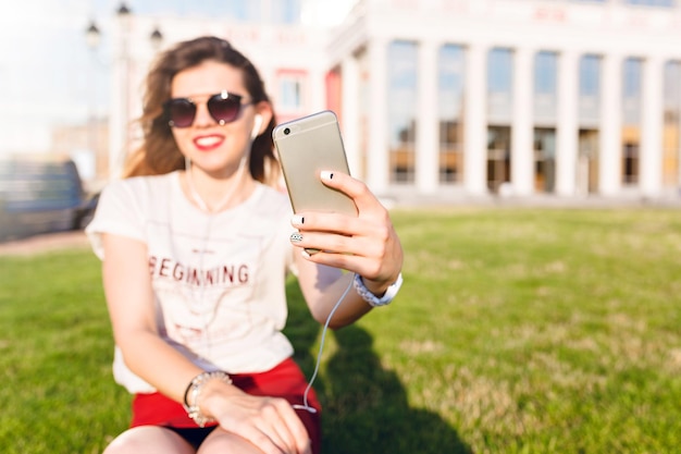 Крупный план смартфона в руках девушки, сидящей на зеленой траве в городском парке. Девушка носит белую футболку, красную юбку и темные солнцезащитные очки. Она делает селфи и широко улыбается.
