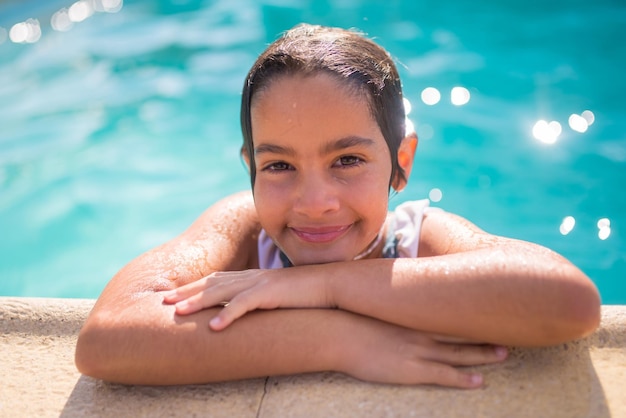 반짝이는 푸른 물 배경에 웃는 소녀의 클로즈업. 수영장에 서 있는 수영복을 입은 백인 소녀는 젖은 손에 턱을 기대고 카메라를 바라보고 있습니다. 활동적인 휴식과 평온한 어린 시절 개념