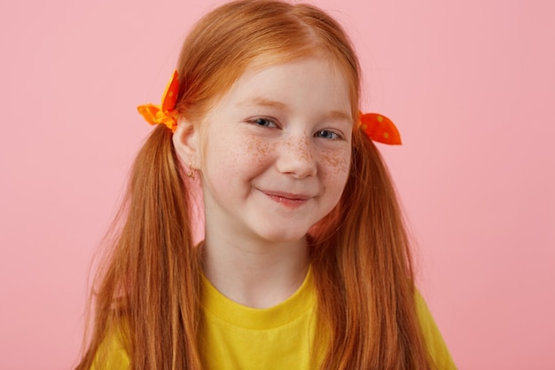 Free photo close up of smiling petite freckles red-haired girl with two tails, smiling and looks cute, wears in yellow t-shirt, stands over pink background.