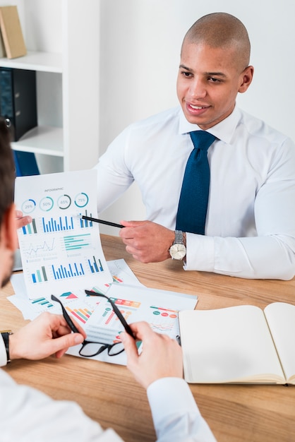 Free photo close-up of a smiling young businessman showing graph with pencil to his business partner at workplace