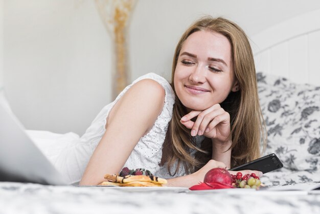 ノートパソコンを見て朝食付きのベッドに横になっている笑顔の若い女性のクローズアップ