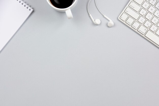 Close-up of spiral notepad; coffee cup; earphone and keyboard on gray background