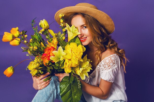 紫の壁の背景の近くのカラフルな春の花の花束を保持しているスタイリッシュな麦わら帽子で美しい金髪の若い女性の春の肖像画を閉じます。