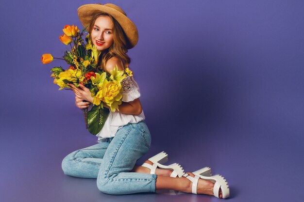 Close up spring portrait of  beautiful blonde young lady in stylish straw summer hat holding   colorful spring flower bouquet   near  purple wall background.