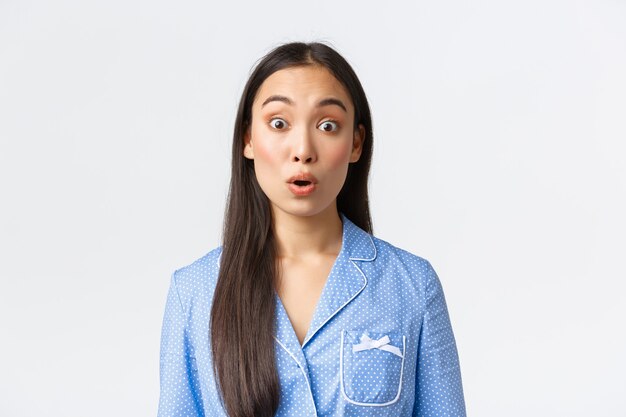 Close-up of startled and shocked asian girl in blue pajama seeing something astounding, look with awe and say wow, staring amazed at camera over white background