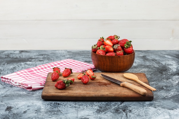 Close-up strawberries and kitchen utensils on wooden cutting board with red gingham tablecloth and a bowl of strawberries on dark blue marble and white wooden background. horizontal