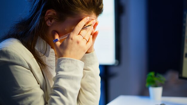 밤늦게 사무실에서 일하는 두통으로 스트레스를 받는 바쁜 여성을 클로즈업하세요. 작업 확인 보고서를 위해 초과 근무를 하는 현대 기술 네트워크 무선을 사용하는 피곤한 집중 직원
