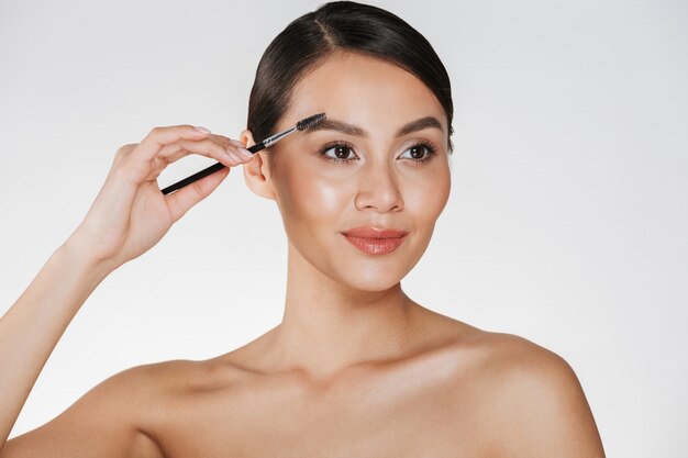 Close up studio portrait of beautiful brunette woman with soft skin painting her eyebrows with makeup brush, isolated over white