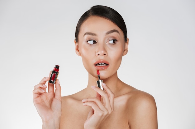 Close up studio portrait of brunette woman with soft skin applying red lipgloss at her lips and looking away, isolated over white