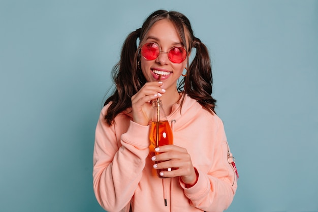 Primo piano ritratto in studio di sognante bella ragazza con i capelli raccolti indossando occhiali rotondi rosa e pullover rosa che beve succo sopra la parete blu isolata