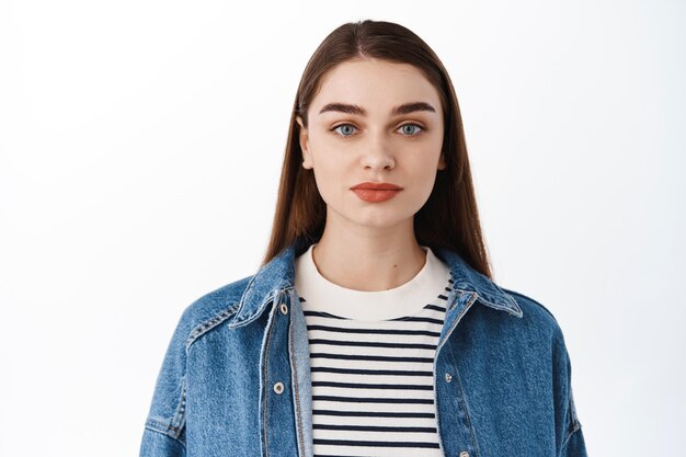 Close up of stylish caucasian girl in denim jacket going on spring walk, looking casual at camera, standing against white background