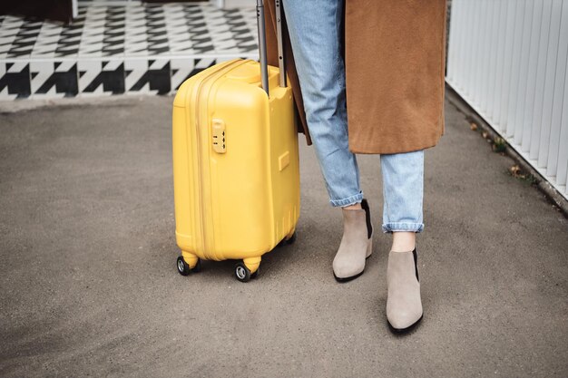 Close up stylish girl in coat standing with yellow suitcase on city street