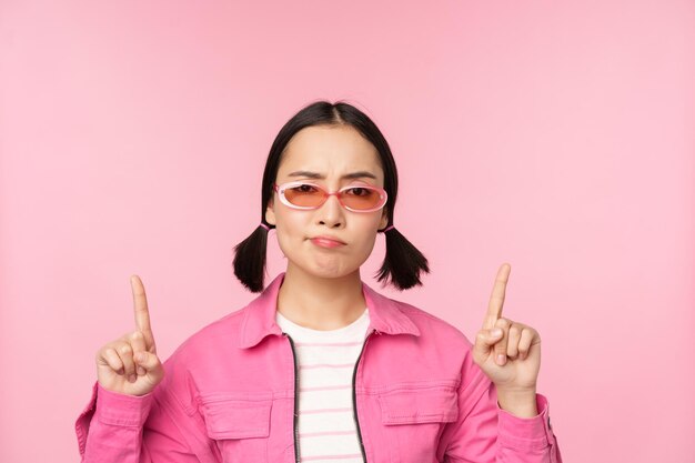 Close up of stylish korean girl looking perplexed pointing fingers up showing advertisement standing over pink background