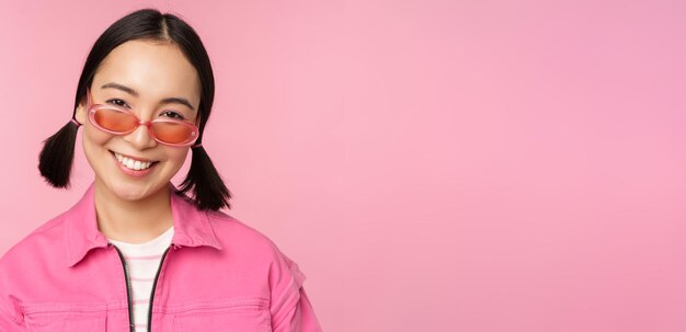 선글라스를 끼고 분홍색 배경에 행복한 포즈를 취하고 있는 세련된 한국 소녀의 클로즈업
