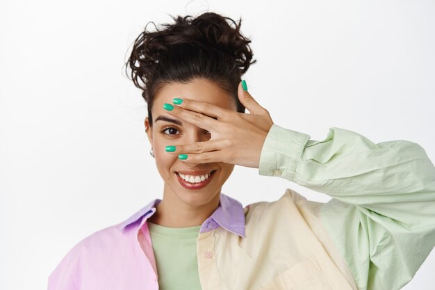 Close up of stylish young modern girl, con eyes, peek through fingers and smiling white teeth, standing in trendy shirt on white