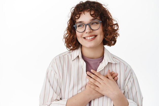 Close up of stylish young woman in glasses looking touched and pleased aside, holding hands on heart and smiling grateful, thanking for something, standing over white background