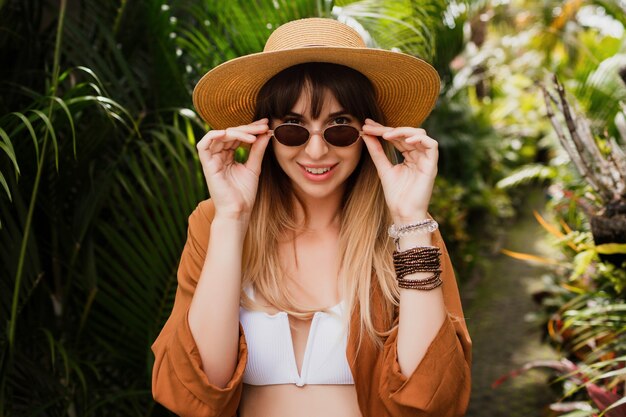 Close up summer fashionable portrait of brunette woman in straw hat posing on tropical palm leaves in Bali.