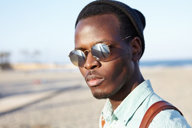 Close up summer outdoor portrait of handsome fashionable Afro American student in mirrored lens sunglasses having walk on beach after college