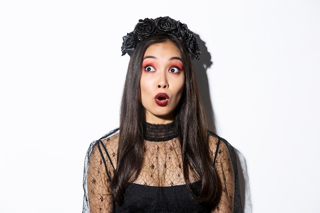 Close-up of surprised asian woman in witch costume looking at upper left corner and open mouth wondered, standing over white background.