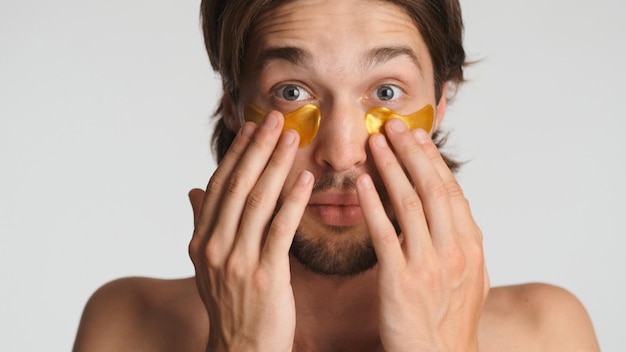 Close up surprised brunette man applying eye patches looking at camera isolated Guy taking care of his skin against a white background