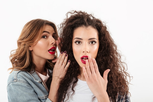 Close up surprised curly girl covering her mouth  while her friend talking her in ear over white wall