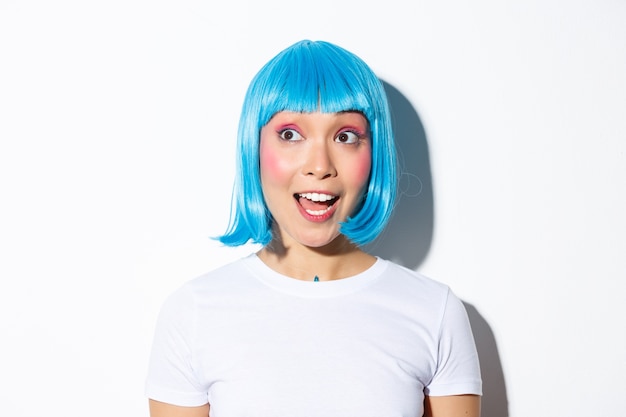 Close-up of surprised cute asian woman in blue wig, looking at halloween banner, standing.