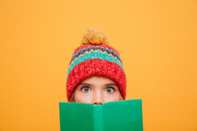 Бесплатное фото Закройте удивлен молодая девушка в свитер и шляпу, прячась за книгу и глядя на камеру над оранжевым
