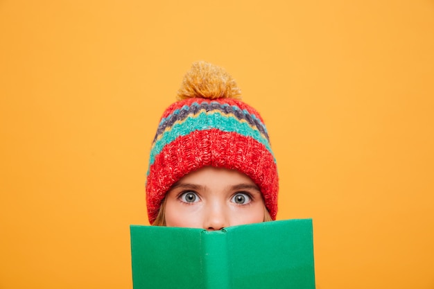 Close up Surprised Young girl in sweater and hat hiding behind the book and looking at the camera over orange