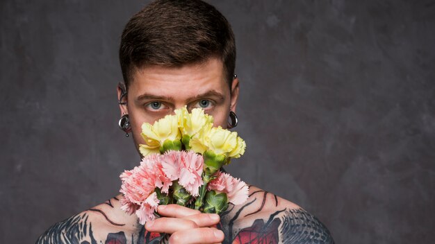 Close-up of tattoo young man with pierced ears holding pink and yellow carnation flowers in front of his mouth