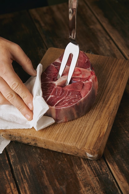 Close up tattooed butcher hand holds meat fork in fresh raw steak for dinner, unrecognizable