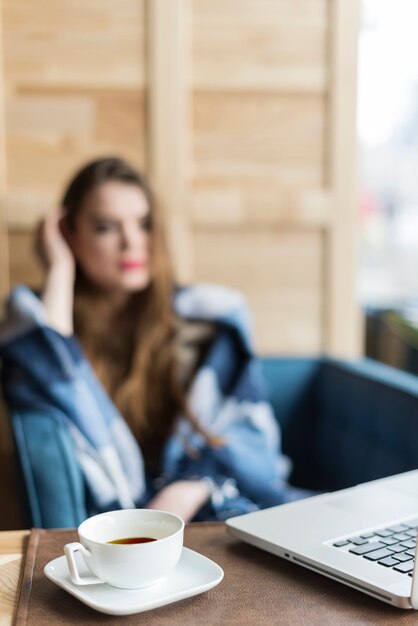 ぼやけた女性の背景とティーカップとラップトップのクローズアップ