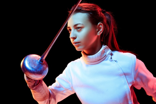 Close up. Teen girl in fencing costume with sword in hand isolated on black background, neon light. Young model practicing and training in motion, action. Copyspace. Sport, youth, healthy lifestyle.