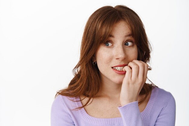 Close up of teenage girl biting finger nails with excitement, looking left eager to try something, feeling tempation, standing in blouse against white background