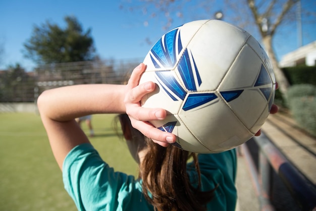 ボールを持つ10代の少女の手のクローズアップ。学校のスタジアムで彼女の頭の上に白と青のボールを持った少女の背面図、彼女の友人にそれを投げる準備ができています。女子サッカー、健康的なライフスタイルのコンセプト