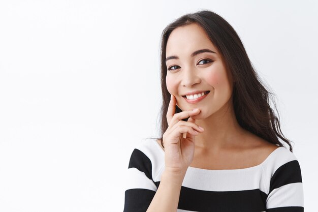 Close-up tender and feminine pretty woman with clean skin no clogged pores, without blemishes, touching cheek and smiling joyfully, gazing camera, tilt head gently, stand white background