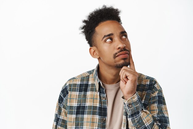 Close up of thoughtful african american man making decision, student thinking about smth, touch jaw and looking up, ponder choices, standing on white